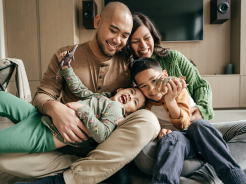 Man and woman gently cuddling and wrestling two children while sitting upright on their living room floor